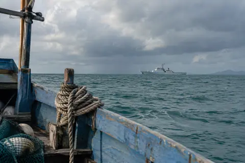 AI-generated illustration: A small fishing boat with a distant patrol vessel under tropical rain clouds.